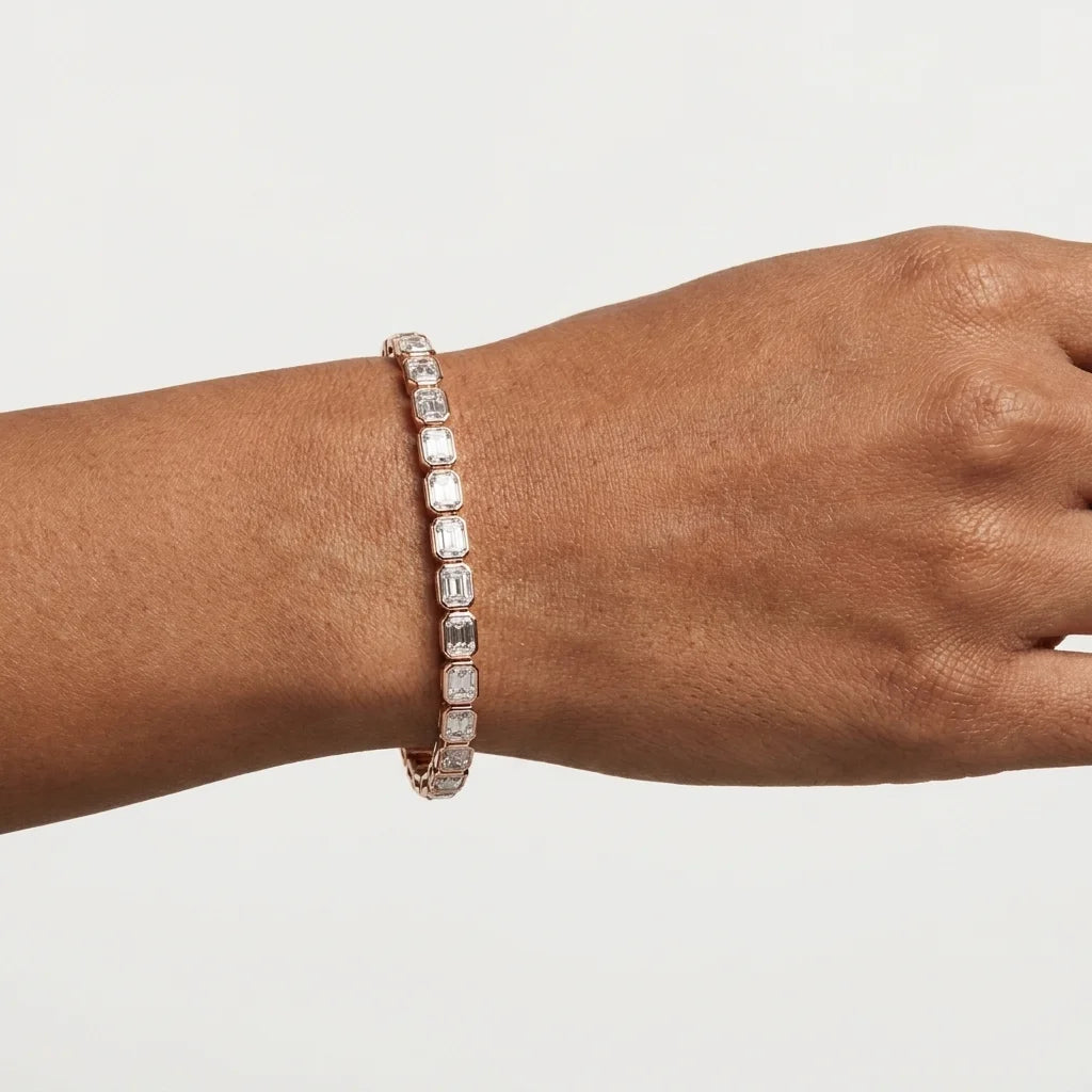A close-up of a person's wrist wearing a slim rose gold tennis bracelet with clear emerald shaped diamonds, set against a plain light gray background.