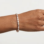 A close-up of a person's wrist wearing a slim rose gold tennis bracelet with clear emerald shaped diamonds, set against a plain light gray background.