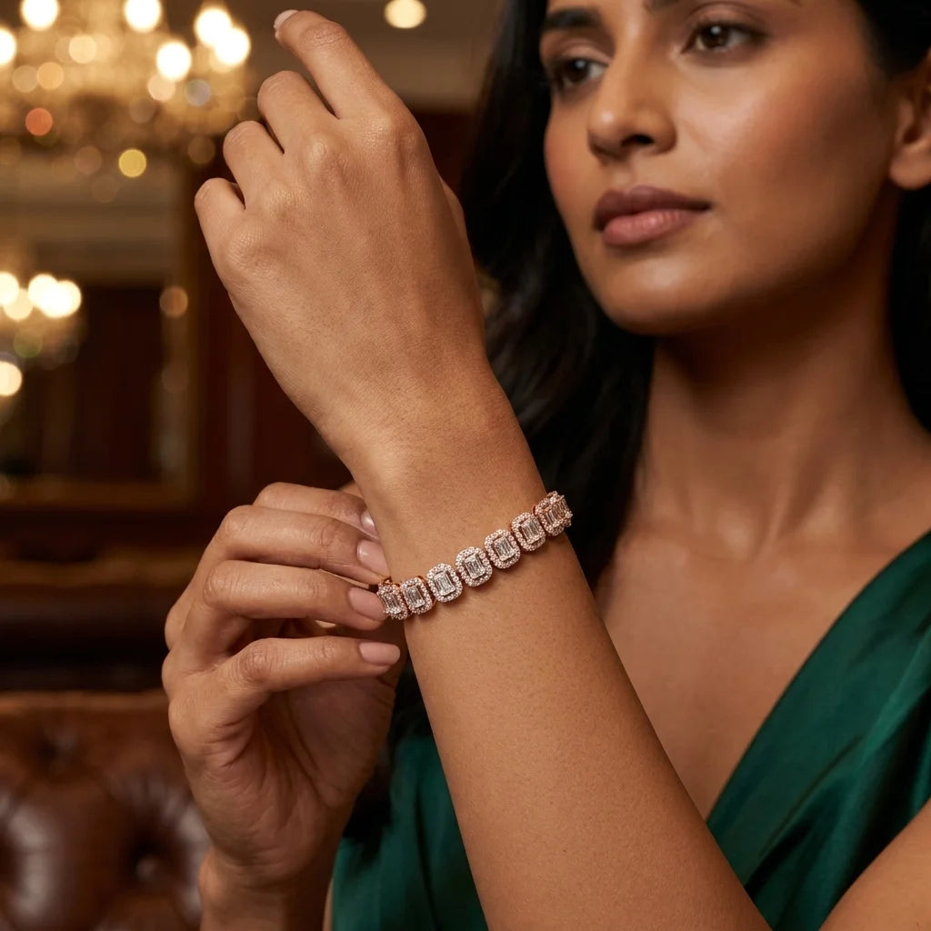 A woman wearing a delicate bracelet with large clear radiant shape diamonds, shown in a warm, elegant indoor setting with blurred chandelier lights in the background.