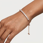 A woman wearing a delicate rose gold tennis bracelet with clear diamonds, shown in a close-up portrait against a dark, elegant background.