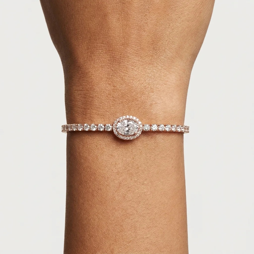 A close-up of a person's wrist wearing a delicate bracelet with a row of clear diamonds and centre of the a large oval diamond halo by small round diamonds.