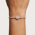 A close-up of a person's wrist wearing a delicate bracelet with a row of clear diamonds and centre of the a large oval diamond halo by small round diamonds.