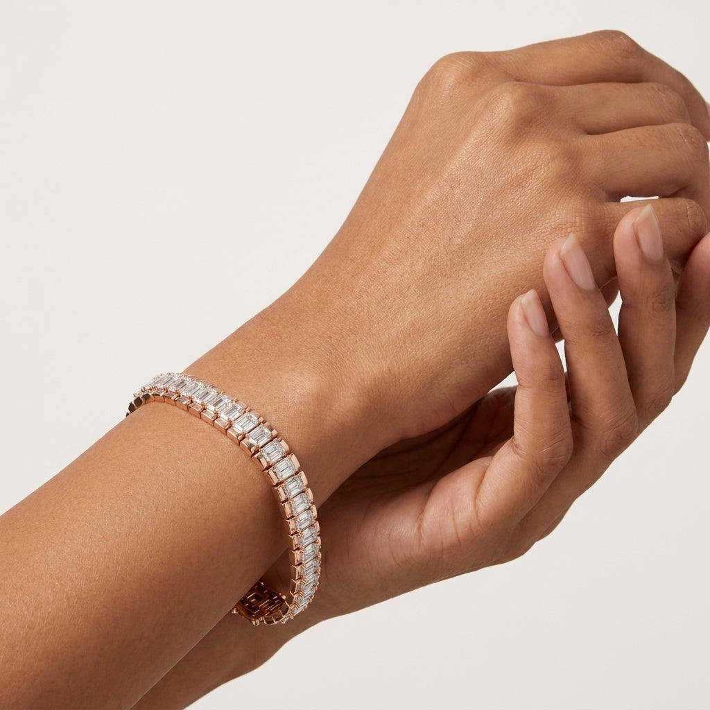 A close-up of a person's wrist and hand wearing a rose gold tennis bracelet with clear emerald-cut diamonds, set against a clean, light off-white background.