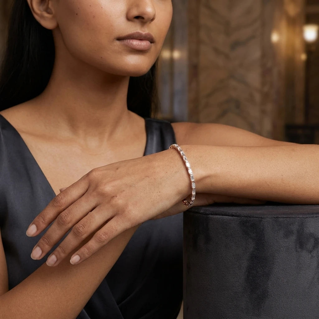A close-up lifestyle shot of a woman wearing a delicate rose gold tennis bracelet with small clear diamonds, resting her arm on a dark velvet surface.