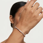 A close-up shot of a woman's hand wearing a delicate rose gold tennis bracelet with small clear diamonds against a white background.