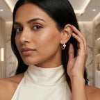 A woman wearing a delicate gold hoop earring with diamonds, shown in a close-up portrait. She is wearing a cream-colored high-neck top and touching her ear with her hand, set against a softly blurred marble-walled interior.