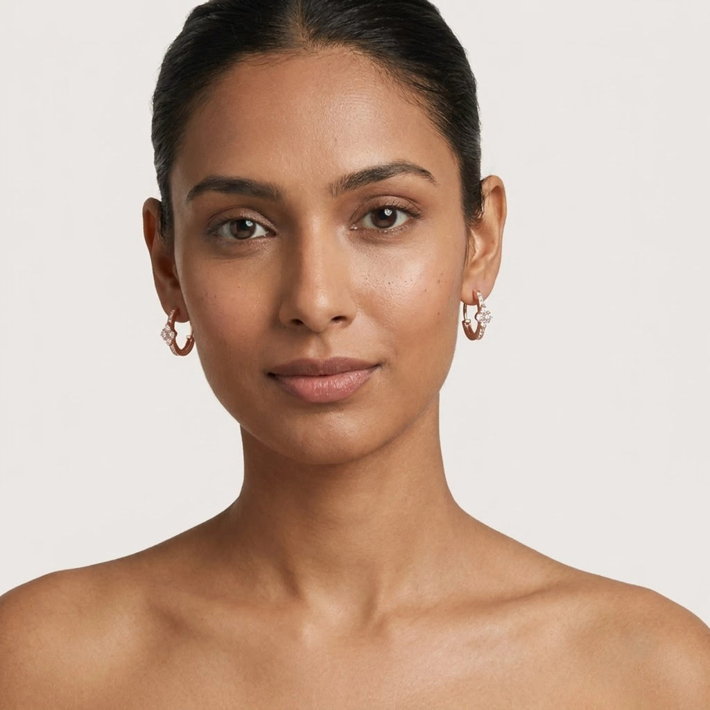 A close-up portrait of a woman wearing small rose gold hoop earrings with clear diamonds details, set against a plain light gray background.