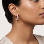 A close-up of a model's ear wearing a rose gold huggie hoop earring adorned with small clear pavé diamonds, set against a clean light gray background.