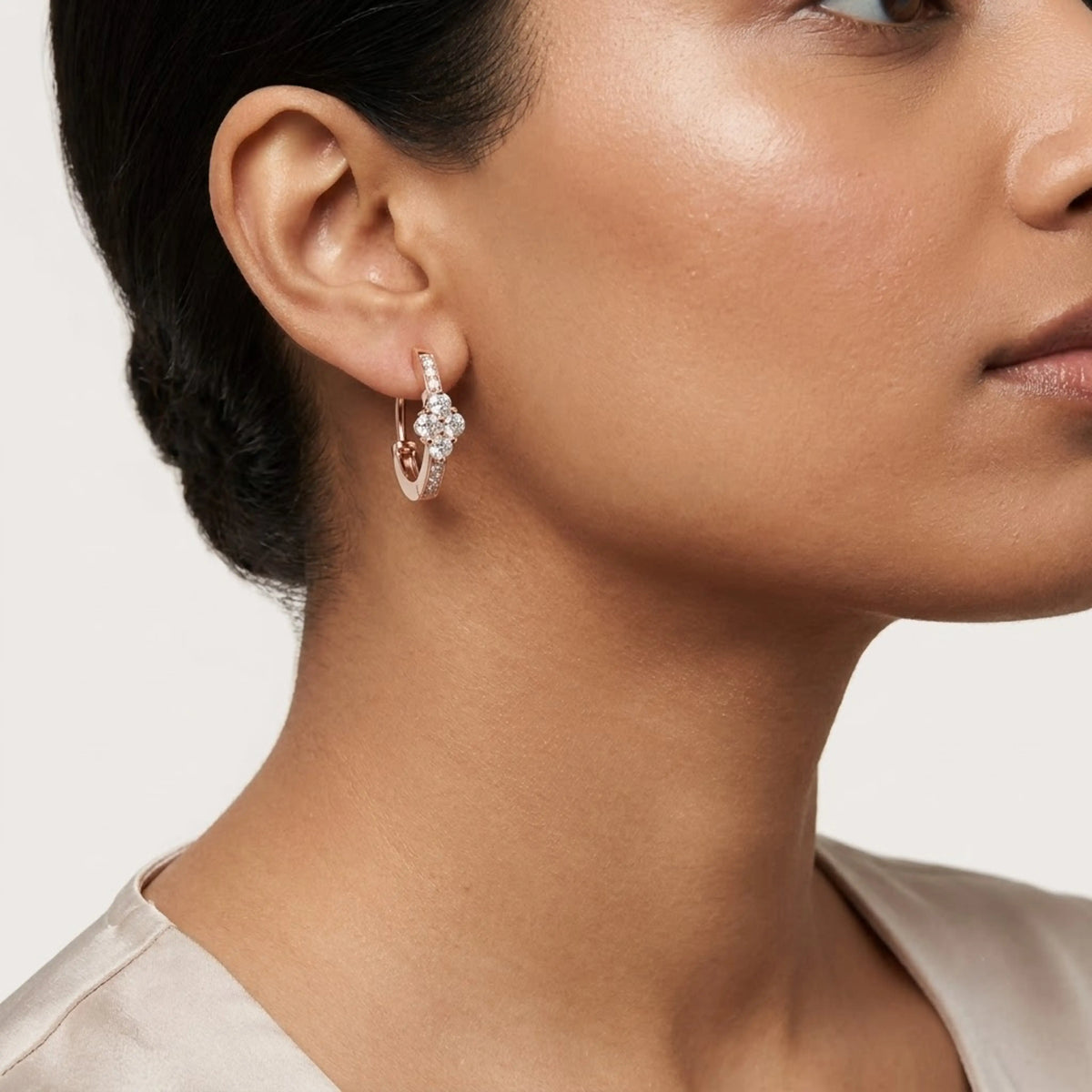 A close-up of a model's ear wearing a rose gold huggie hoop earring adorned with small clear pavé diamonds, set against a clean light gray background.