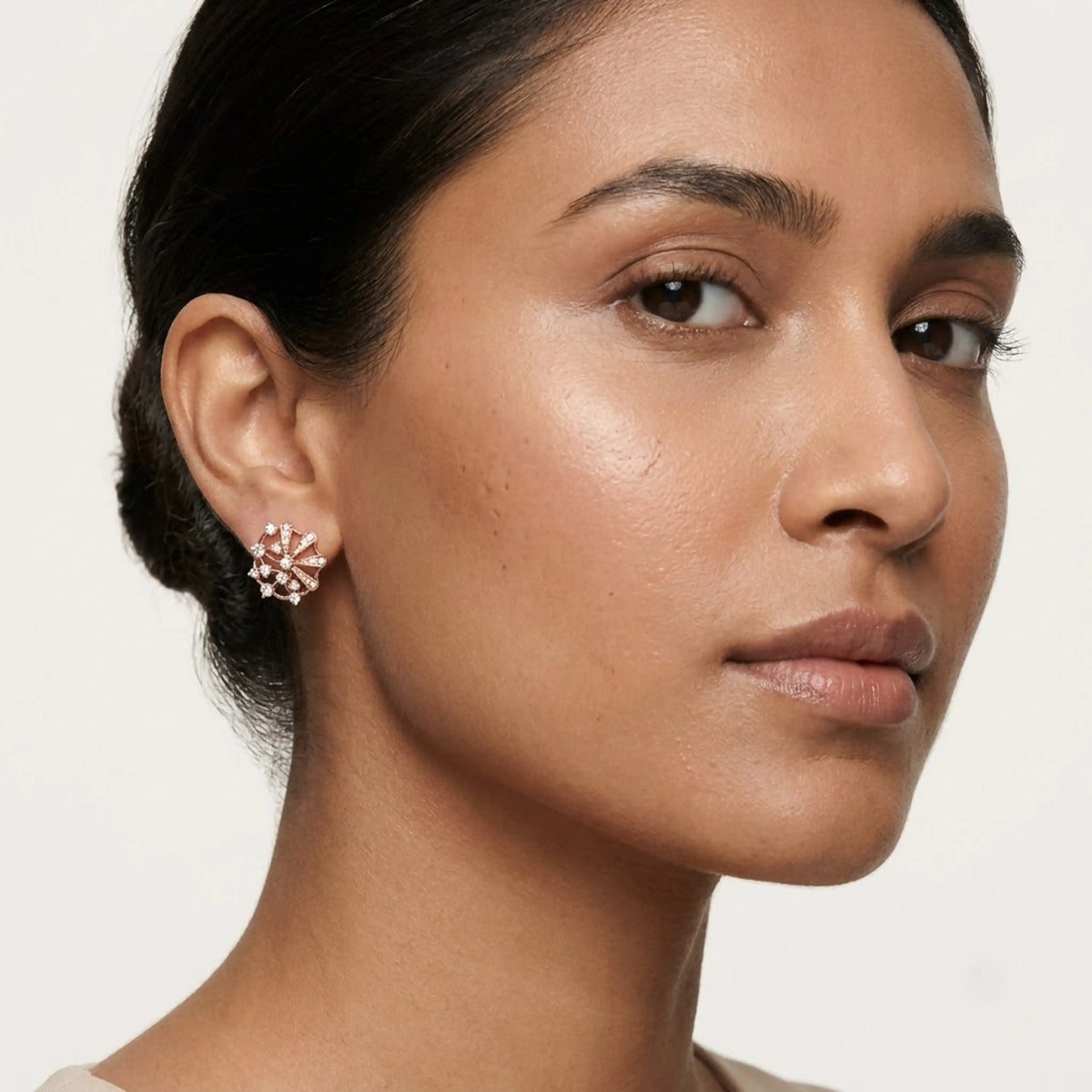 A close-up of a woman wearing a delicate rose gold earring with small clear stones, set against a clean, light off-white background.