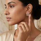 A close-up side profile of a woman wearing a single round rose gold stud earring with clear diamonds, touching her neck to show the earring. She is wearing a light beige satin top, and the background is a softly blurred warm-toned interior.