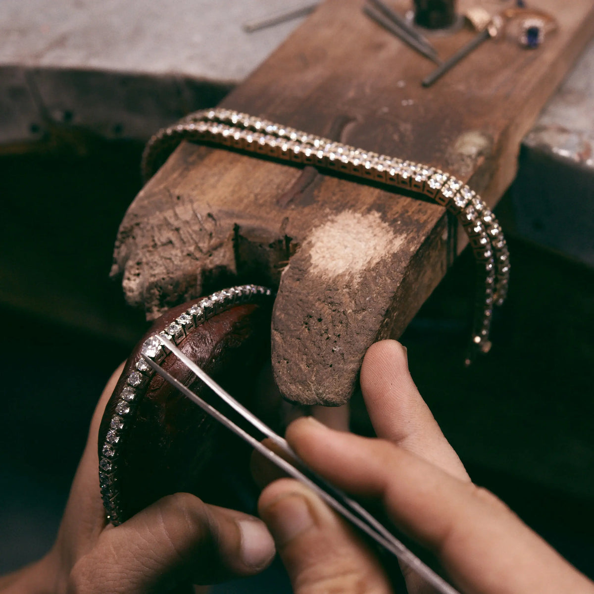 A close-up of hands using tweezers to place a clear diamonds bracelet onto a wooden jeweler's bench, with two matching bracelets resting on the wood.