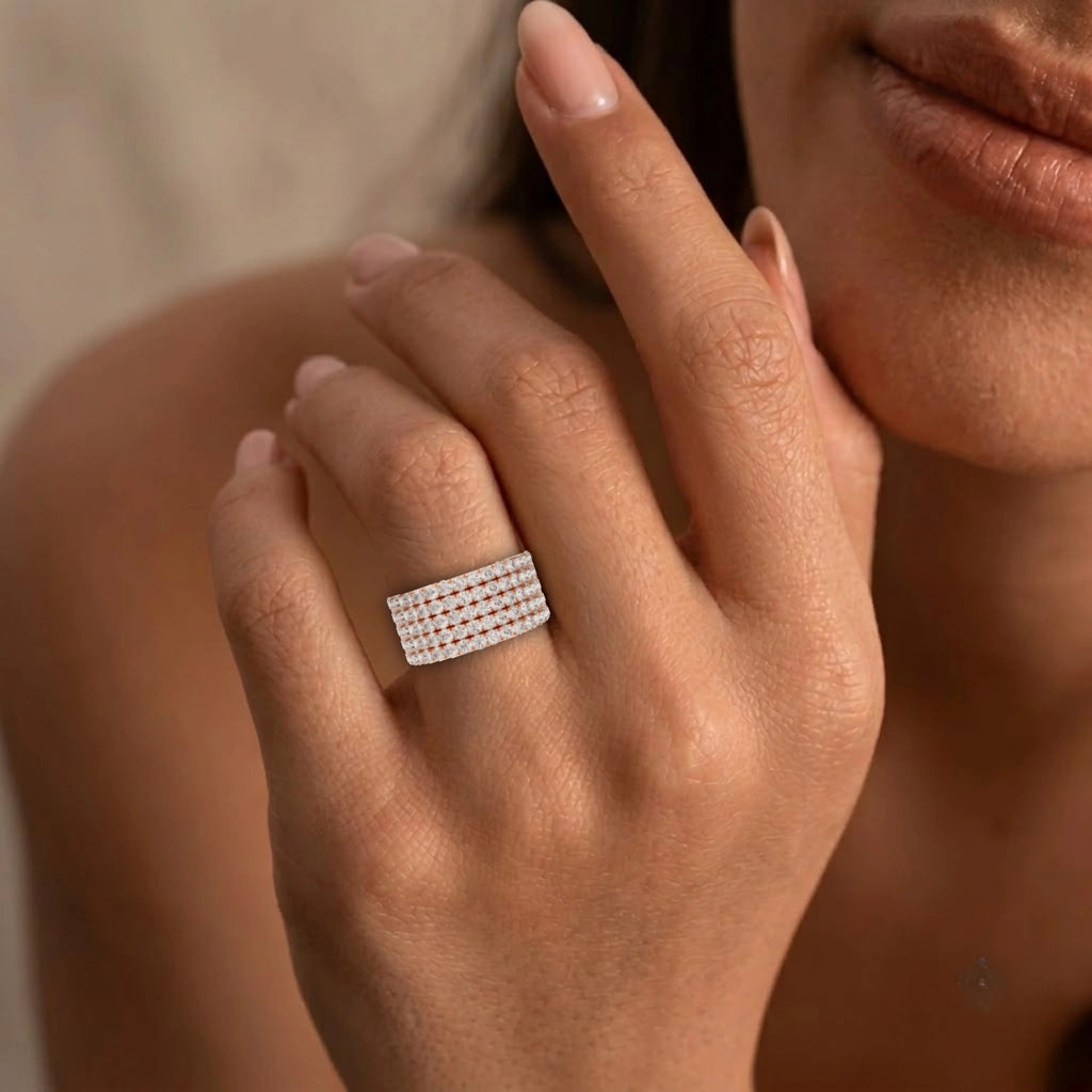 A close-up of a woman's hand wearing a wide band ring paved with small clear stones, against a softly blurred warm beige background.