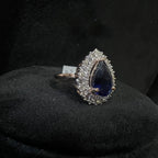 Ring with a teardrop-shaped blue gemstone and diamond-studded halo on a black background