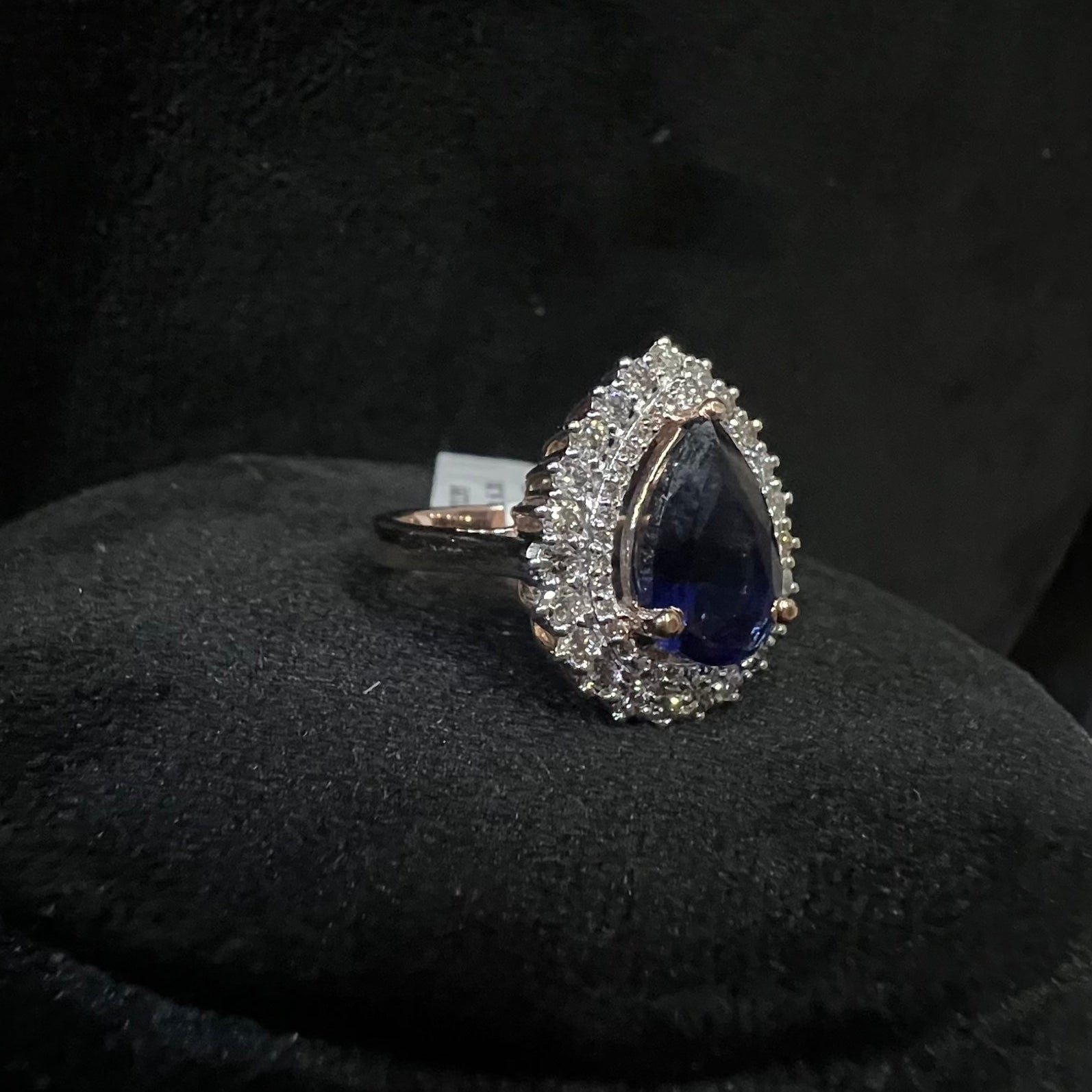 Ring with a teardrop-shaped blue gemstone and diamond-studded halo on a black background