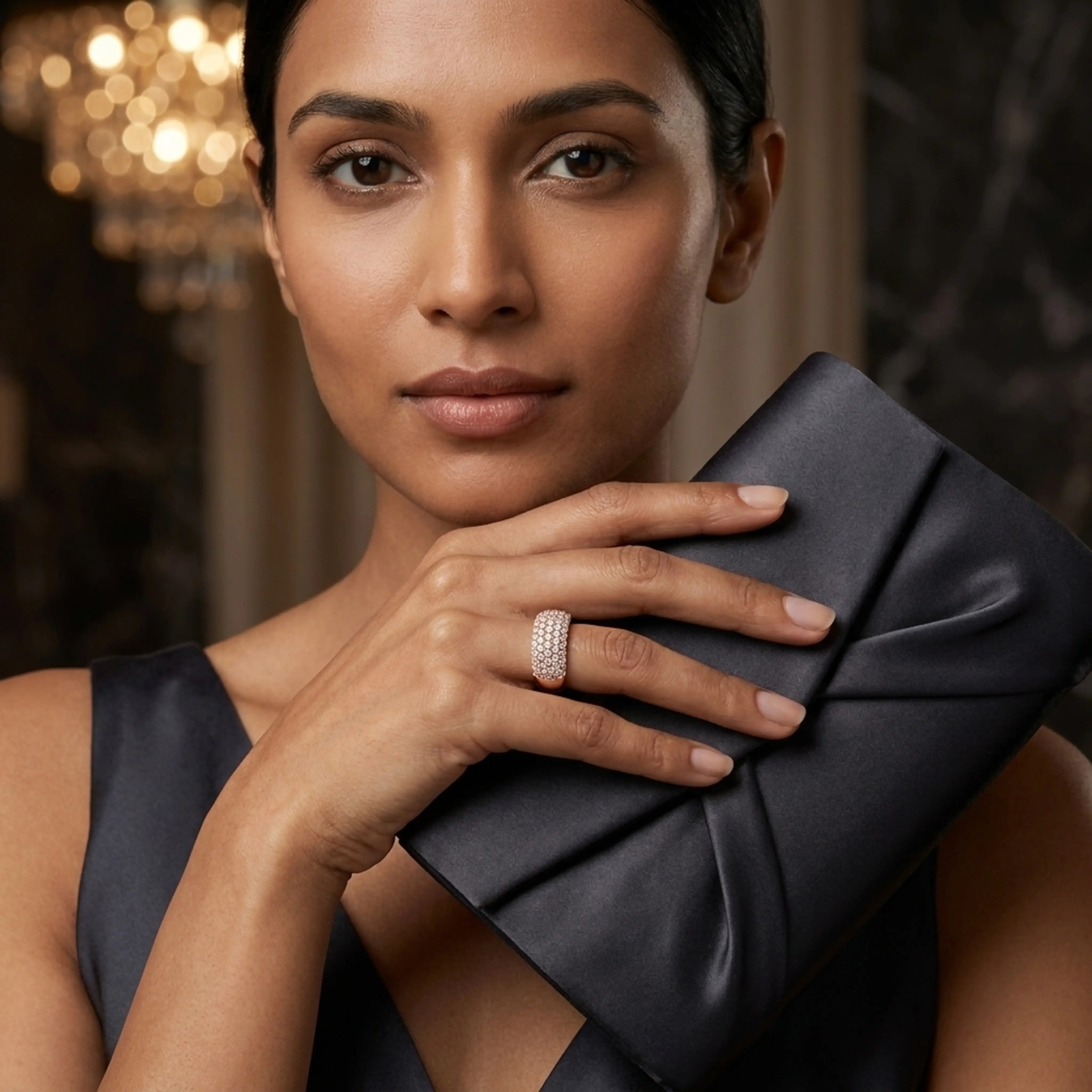 A close-up lifestyle shot of a woman wearing a sparkling diamond band ring, holding a dark gray satin clutch in a luxurious indoor setting with a blurred chandelier in the background.